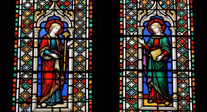 Detail of a Gothic stained glass window showing figures against a patterned blue and red glass background, light streaming through.