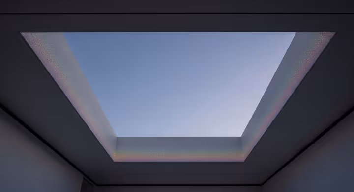 An interior view of a James Turrell Skyspace, where viewers look up through an opening in the ceiling at the sky, framed by shifting colored lights.