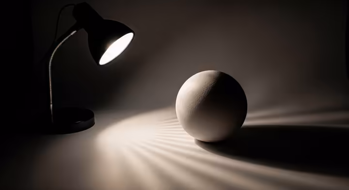 A simple still life setup for a lighting exercise: a matte white sphere on a gray surface, lit from the side by a desk lamp in a dark room.